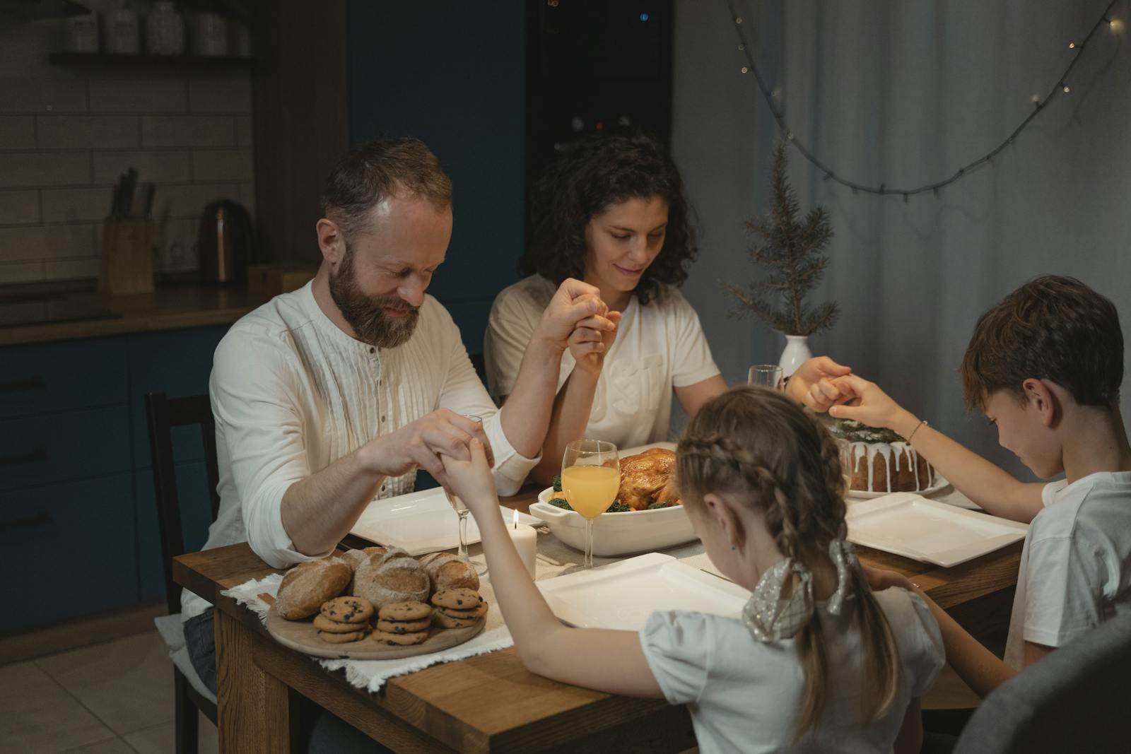 Family holding hands and praying over a dinner table with turkey and cookies during festival celebration.